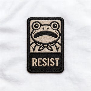 May include: Embroidered patch featuring a cartoon frog with large eyes and a bow tie. The frog is set against a tan background, with the word "RESIST" in white block letters below. The patch has a black border.