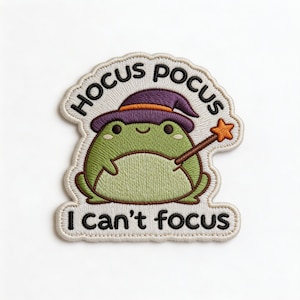 May include: Embroidered patch featuring a green frog wearing a purple witch hat and holding a star-tipped wand. The patch reads "Hocus Pocus" above the frog and "I can't focus" below, all in black lettering.