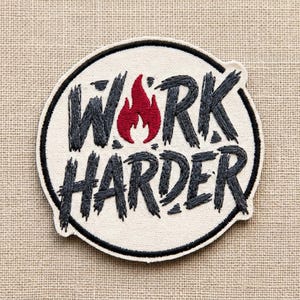 May include: Embroidered patch with the words "WORK HARDER" in dark gray thread, with a red flame graphic. The patch is circular, with a black border and a white background. The patch is on a tan fabric.