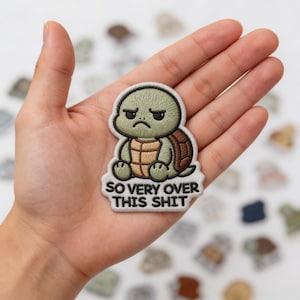 May include: Embroidered patch featuring a cartoon turtle with a grumpy expression. The turtle has a green head and brown shell. The text "SO VERY OVER THIS SHIT" is below the turtle in white letters. The patch is held in a hand.