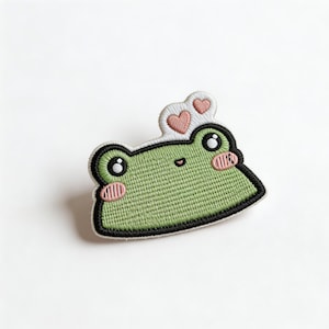 May include: Embroidered patch featuring a cartoon frog with a light green body, black outline, and pink cheeks. Two pink hearts sit above the frog's head. The patch is designed with a textured, stitched appearance, perfect for adding to clothing or accessories.