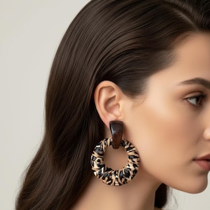May include: A close-up of a person wearing a pair of large hoop earrings. The earrings have a dark brown rectangular top and a circular hoop with a black and tan animal print design. The background is a solid light color.