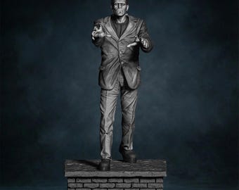Op de afbeelding: Een grijswaarden sculptuur van Frankenstein's monster, gekleed in een pak en staand op een bakstenen platform. De figuur heeft een serieuze uitdrukking en uitgestrekte armen, tegen een donkerblauwe achtergrond.