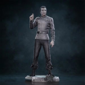 May include: A gray figurine of a man in a uniform, with a belt buckle featuring a circular emblem. The figure is standing on a detailed base. The man is raising his right hand with his index finger extended.