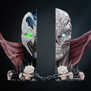 May include: Spawn bookends, split to show the character's face. One side has a white mask with green eyes, the other a skeletal face with glowing blue eyes. Both sides have a red cape and are decorated with skulls. A chain connects the two halves.