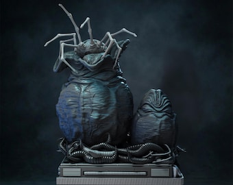 Op de afbeelding: Een gedetailleerde sculptuur van een eierzak van een wezen en een ei, met een spinnenachtig gezicht dat uit de top komt. De sculptuur is donkerblauw en grijs, met ingewikkelde details en een getextureerd oppervlak. De basis is metallic grijs.