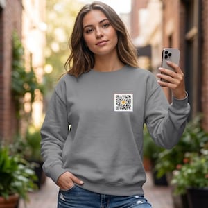 May include: A grey sweatshirt with a small square patch featuring a QR code with a yellow and black design. The sweatshirt has long sleeves and a crew neck. The person is holding a smartphone.