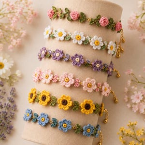 Puede incluir: Exhibición de seis pulseras de flores de ganchillo en varios colores, incluyendo rosa, blanco, amarillo, azul y morado. Cada pulsera presenta un diseño floral diferente con hojas verdes y detalles de cadena dorada.