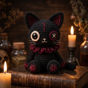 May include: A black crocheted cat doll with a dark red ruffled collar, one large glossy eye, and one button eye with an X-stitch. The cat features a stitched forehead detail, dark red accents on the ears and paws, and white embroidered whiskers.
