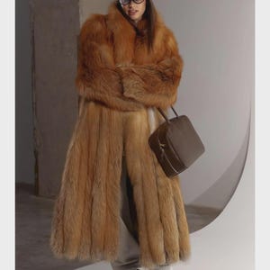 Puede incluir: Un abrigo de piel largo y grueso de color óxido, combinado con una gorra de béisbol marrón, gafas de montura oscura y un bolso de cuero marrón estructurado. El conjunto incluye pantalones de chándal claros, calcetines blancos y zapatillas beige con cordones verdes.