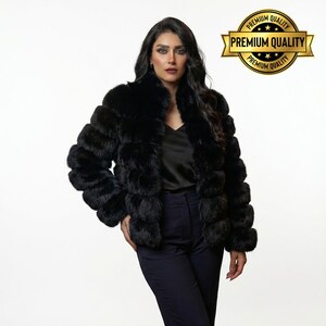 May include: A black faux fur jacket with a layered design. The jacket has a high collar and long sleeves. The model is wearing a black top and dark blue pants. A gold emblem with the words "Premium Quality" is in the upper right corner.