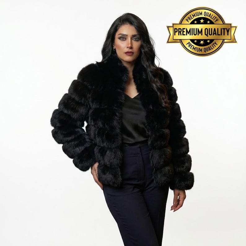May include: A black faux fur jacket with a layered design. The jacket has a high collar and long sleeves. The model is wearing a black top and dark blue pants. A gold emblem with the words "Premium Quality" is in the upper right corner.