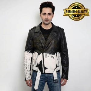 Könnte beinhalten: Eine schwarze Motorradjacke aus Leder mit einem weiß bemalten unteren Bereich, der handschriftlichen Text und Sternmotive aufweist. Die Jacke verfügt über ein Revers mit Metallnieten, einen Frontreißverschluss und einen weißen Gürtel mit Schnalle.