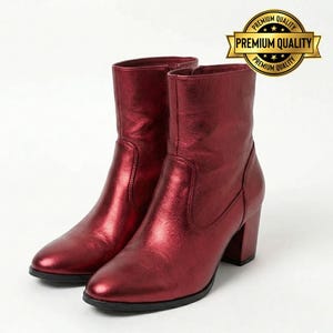 May include: A pair of burgundy-colored ankle boots with a metallic sheen. The boots have a rounded toe, a block heel, and a mid-calf shaft. The boots are displayed on a white background. A gold-colored "Premium Quality" seal is in the upper right corner.