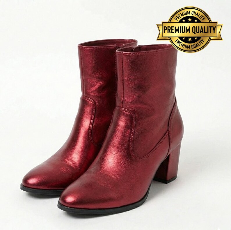 May include: A pair of burgundy-colored ankle boots with a metallic sheen. The boots have a rounded toe, a block heel, and a mid-calf shaft. The boots are displayed on a white background. A gold-colored "Premium Quality" seal is in the upper right corner.