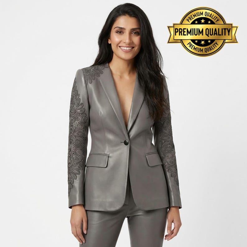 Pode incluir: Um blazer de couro cinza com bordados pretos intrincados nas mangas. O blazer tem um fecho de um bot&atilde;o e dois bolsos frontais. Cal&ccedil;as cinza combinando tamb&eacute;m s&atilde;o vis&iacute;veis. A imagem inclui um emblema dourado com o texto "PREMIUM QUALITY".