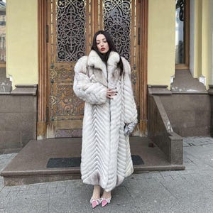 May include: A full-length, white fur coat with a chevron pattern. The coat has a large collar and long sleeves. The person is wearing pink heels with bows and carrying a silver clutch. The background features a wooden door and a building exterior.