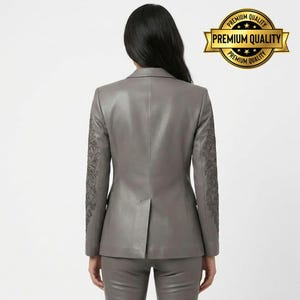 Pode incluir: Um blazer de couro cinza com detalhes bordados intrincados nas mangas. O blazer faz parte de um conjunto a condizer. A imagem inclui um selo de "Premium Quality".