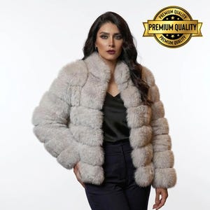 May include: A light gray faux fur jacket with a collar. The jacket has a horizontal design and is worn over a black top and dark pants. A gold seal with the words "Premium Quality" is in the upper right corner.