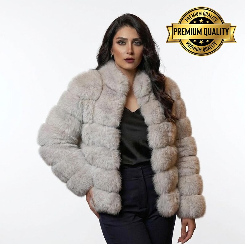 May include: A light gray faux fur jacket with a collar. The jacket has a horizontal design and is worn over a black top and dark pants. A gold seal with the words "Premium Quality" is in the upper right corner.