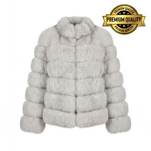 May include: A light gray faux fur jacket with a high collar and horizontal panel design. The jacket has long sleeves and a soft, textured appearance. A gold seal with the words "Premium Quality" is in the upper right corner.