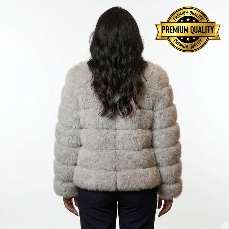 May include: A light gray, horizontally-paneled faux fur jacket. The jacket is shown from the back, with a woman wearing it. A gold seal with the words "Premium Quality" is in the upper right corner.