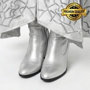 May include: A pair of silver ankle boots with a pointed toe and a small heel. The boots are made of a shiny material and have a black sole. The boots are under a silver garment with a decorative pattern. A gold seal with the text "Premium Quality" is visible.