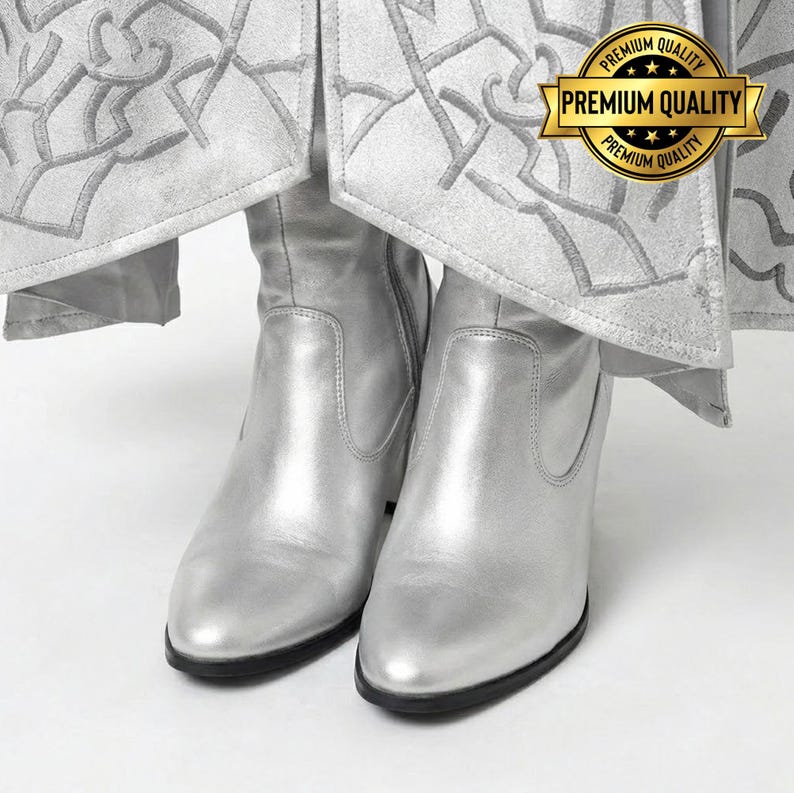 May include: A pair of silver ankle boots with a pointed toe and a small heel. The boots are made of a shiny material and have a black sole. The boots are under a silver garment with a decorative pattern. A gold seal with the text "Premium Quality" is visible.