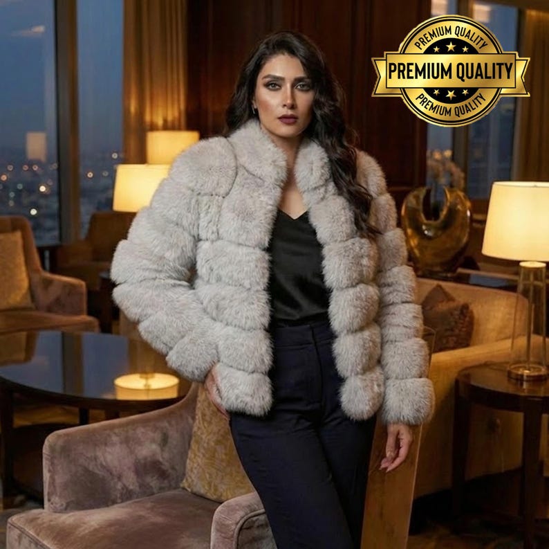 May include: A light gray fur jacket with a wide collar and horizontal panel design. The jacket is worn over a black top and dark pants. A gold-colored "Premium Quality" seal is in the upper right corner.