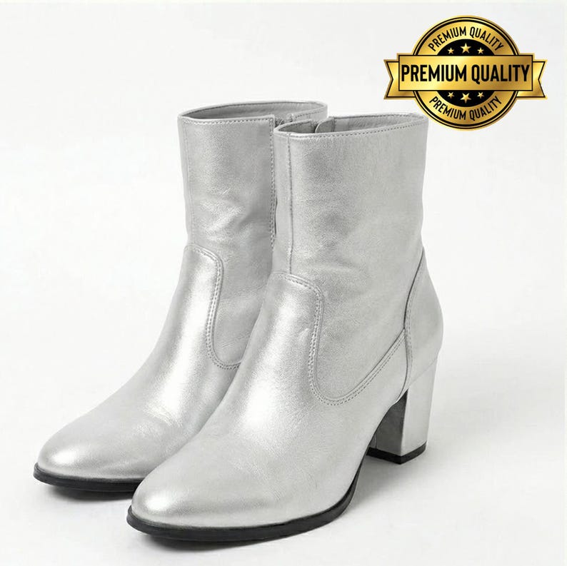 May include: A pair of silver ankle boots with block heels. The boots have a shiny, metallic finish and a zipper on the side. The boots are displayed on a white background. A gold seal with the words "Premium Quality" is in the upper right corner.