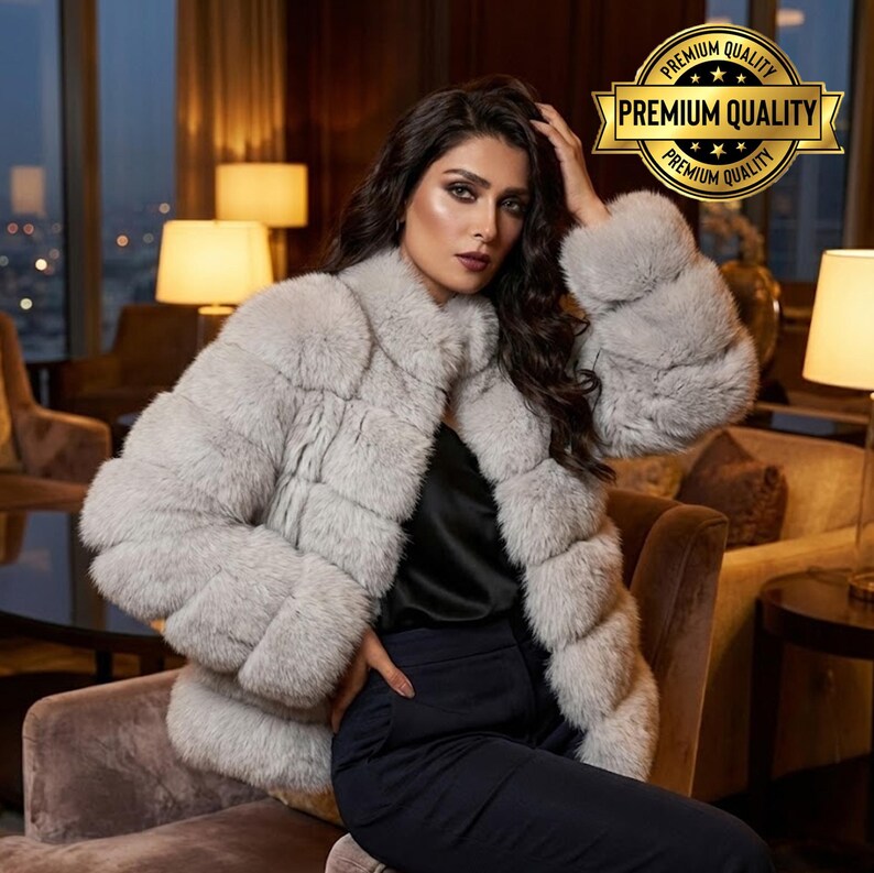 May include: A light gray fur coat with a textured design. The coat has a round collar and long sleeves. The image also includes a black top and dark blue pants. A gold seal with the words "Premium Quality" is in the upper right corner.