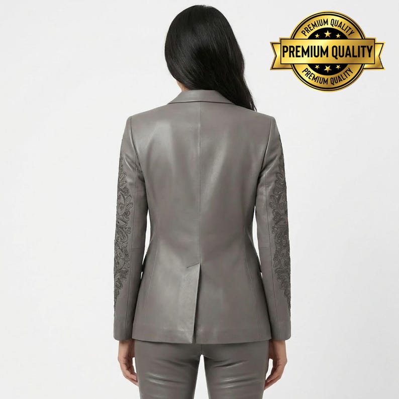 Pode incluir: Um blazer de couro cinza com detalhes bordados intrincados nas mangas. O blazer faz parte de um conjunto a condizer. A imagem inclui um selo de "Premium Quality".