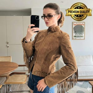 May include: A brown suede jacket with fringe detailing on the sleeves. The jacket has a zip-up front and a fitted silhouette. The image also includes a phone, sunglasses, and a "Premium Quality" seal.