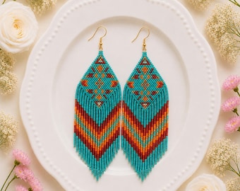 Könnte beinhalten: Ein Paar lange Ohrhänger aus winzigen Saatperlen in Türkis, Orange und Dunkelrot. Die Ohrringe zeigen ein geometrisches Rautenmuster im oberen Bereich und einen Fransenabschluss im Chevron-Stil, befestigt an goldfarbenen Ohrhaken.