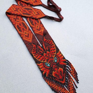 May include: A beaded dragon belt in shades of red, orange, and black. The dragon's head is at the bottom, with turquoise eyes and a fringed beard. The belt's strap features geometric patterns.