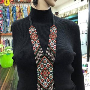 Op de afbeelding: Een kralenketting met een geometrisch ontwerp in rood, groen en wit. De ketting heeft een zwarte achtergrond en een franje aan de onderkant. Zwarte kralen oorbellen zijn ook zichtbaar. De ketting wordt op een mannequin getoond.
