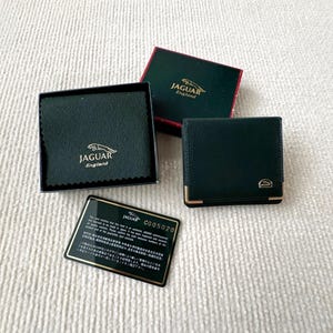 Op de afbeelding: Een donkergroene lederen portemonnee met goudkleurige hoekaccenten en een klein metalen logo, vergezeld van een bijpassende doos, een beschermende doek met het Jaguar England-logo en een zwarte echtheidskaart met het serienummer C005020.