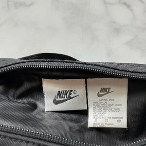 Pu&ograve; includere: Primo piano di una borsa in nylon nera con due etichette bianche. Le etichette mostrano il logo Nike e le informazioni sul prodotto, tra cui "S40710 PRS" e le istruzioni per la cura. La borsa ha una cerniera e sembra essere prodotta a Taiwan.