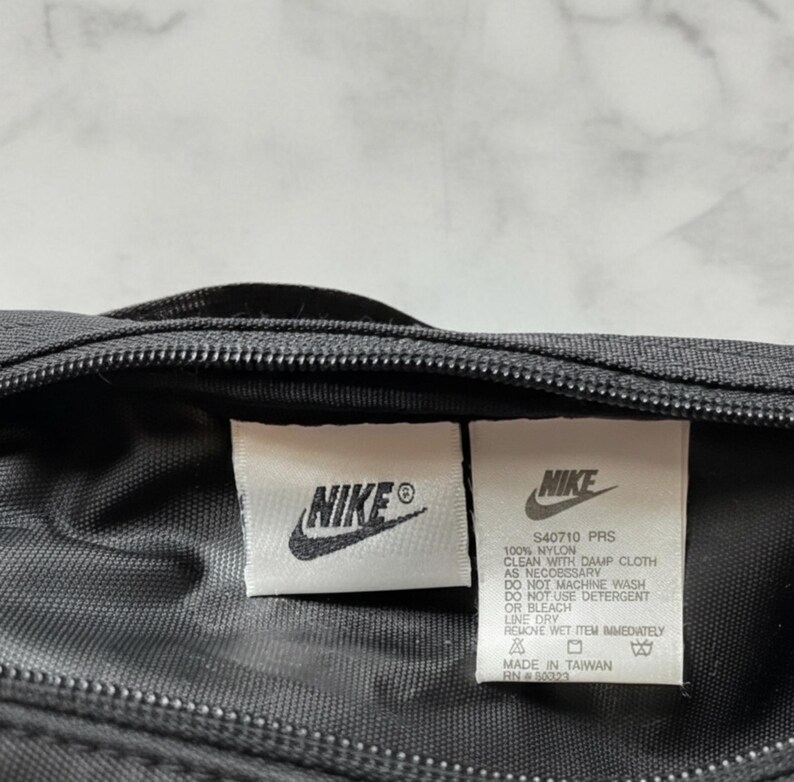 Pu&ograve; includere: Primo piano di una borsa in nylon nera con due etichette bianche. Le etichette mostrano il logo Nike e le informazioni sul prodotto, tra cui "S40710 PRS" e le istruzioni per la cura. La borsa ha una cerniera e sembra essere prodotta a Taiwan.