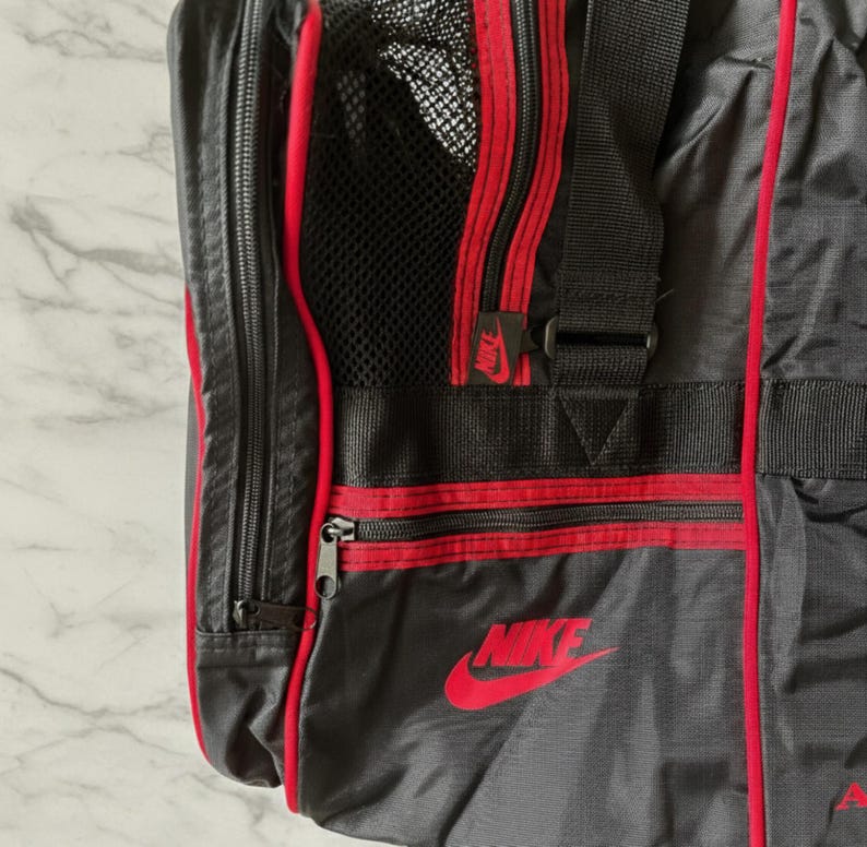 Pu&ograve; includere: Borsa da viaggio Nike nera e grigia con dettagli rossi. Presenta una tasca a rete, cerniere rosse e un logo Nike rosso. La borsa ha cinghie nere e una forma rettangolare.