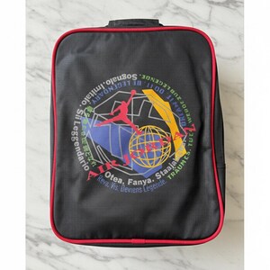 Pu&ograve; includere: Zaino rettangolare nero con bordatura rossa. La parte anteriore presenta una grafica colorata con una silhouette di giocatore di basket, un globo e testo in pi&ugrave; lingue, tra cui "Dream It. Do It. Be Legendary."