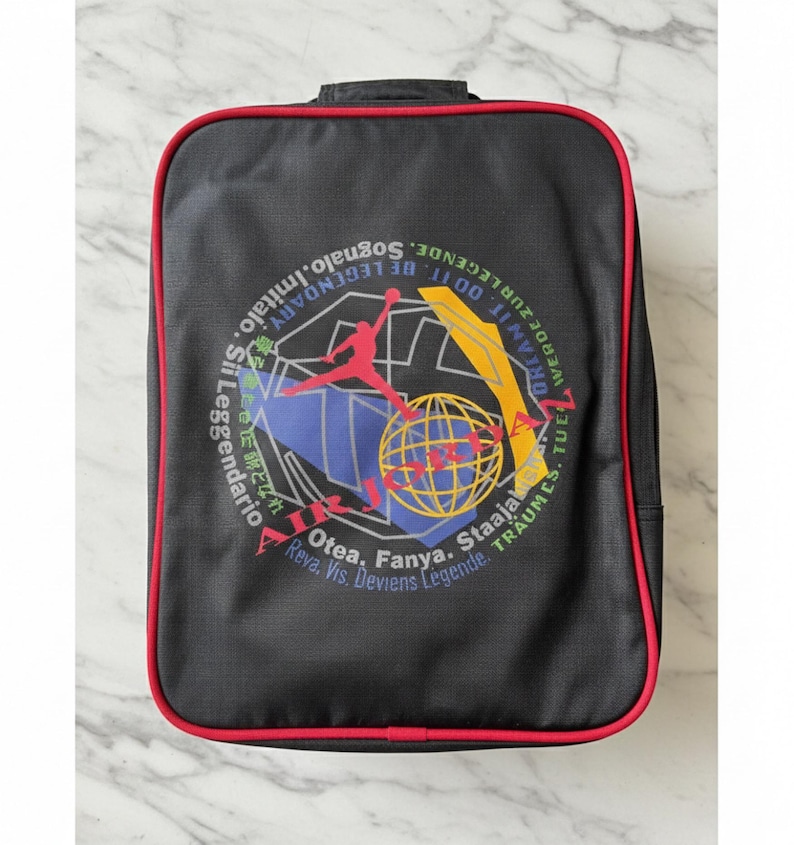 Pu&ograve; includere: Zaino rettangolare nero con bordatura rossa. La parte anteriore presenta una grafica colorata con una silhouette di giocatore di basket, un globo e testo in pi&ugrave; lingue, tra cui "Dream It. Do It. Be Legendary."