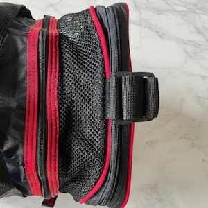 Pu&ograve; includere: Borsa nera con finiture e cerniere rosse. La borsa presenta una tasca a rete e una fibbia nera. Le cerniere e le finiture sono di colore rosso vivo, in contrasto con il tessuto nero. La borsa sembra essere realizzata in un materiale resistente.