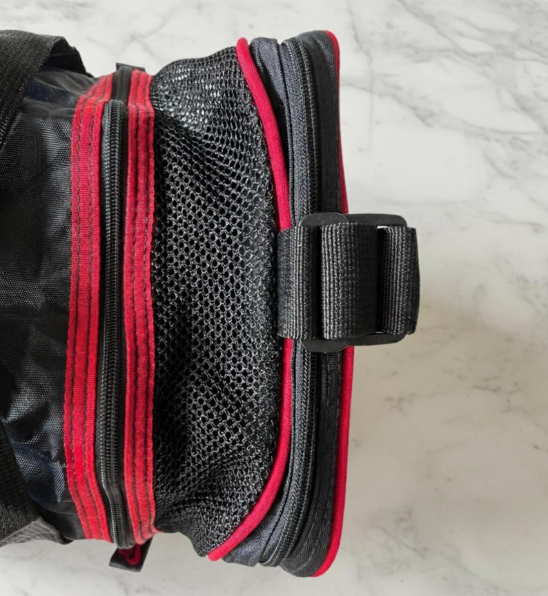 Pu&ograve; includere: Borsa nera con finiture e cerniere rosse. La borsa presenta una tasca a rete e una fibbia nera. Le cerniere e le finiture sono di colore rosso vivo, in contrasto con il tessuto nero. La borsa sembra essere realizzata in un materiale resistente.