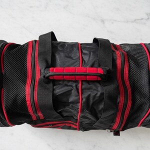 Pu&ograve; includere: Borsa da viaggio nera e rossa con un pannello laterale in rete. La borsa ha due manici neri e una maniglia imbottita rossa al centro. Strisce rosse corrono lungo i lati. La borsa &egrave; rettangolare e ha una chiusura a cerniera.