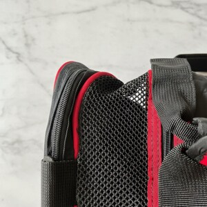 Pu&ograve; includere: Primo piano di un trasportino nero per animali domestici con finiture rosse. Il trasportino presenta un pannello in rete per la ventilazione e una chiusura a zip. Il trasportino ha cinghie nere e un accento rosso lungo i bordi.