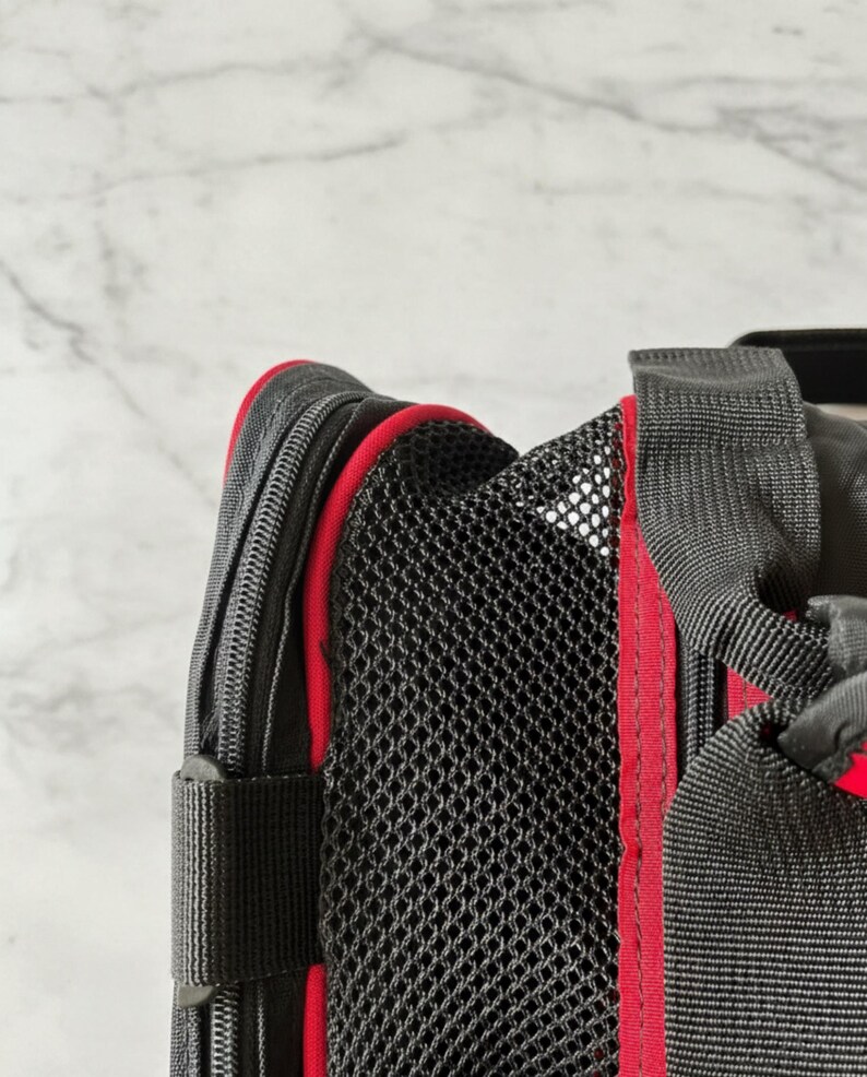 Pu&ograve; includere: Primo piano di un trasportino nero per animali domestici con finiture rosse. Il trasportino presenta un pannello in rete per la ventilazione e una chiusura a zip. Il trasportino ha cinghie nere e un accento rosso lungo i bordi.