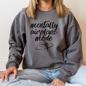 Może przedstawiać: Szara bluza z napisem "mentally airplane mode" w kursywie. Poniżej tekstu znajduje się grafika samolotu ze słowem "ON" w okręgu. Bluza jest w jednolitym kolorze i ma długie rękawy.