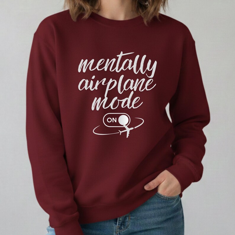 Może przedstawiać: Bordowa bluza z okrągłym dekoltem z białym napisem "mentally airplane mode on". Projekt zawiera stylizowaną ikonę samolotu. Bluza jest w jednolitym kolorze i wydaje się być wykonana z miękkiego materiału.