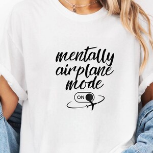 Może przedstawiać: Biała koszulka z czarnym napisem "mentally airplane mode". Poniżej znajduje się grafika samolotu z napisem "ON" w okręgu.